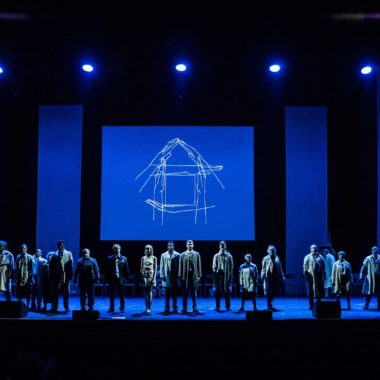a long shot taken of a group of people standing on a stage taking a bow. The stage is lit with blue lighting.