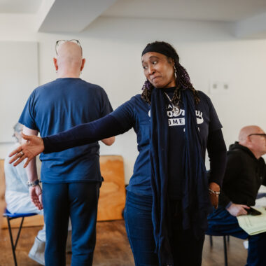A woman gestures with her left hand while looking over her shoulder. She wears a dark blue t-shirt with white lettering and a dark blue scarf.