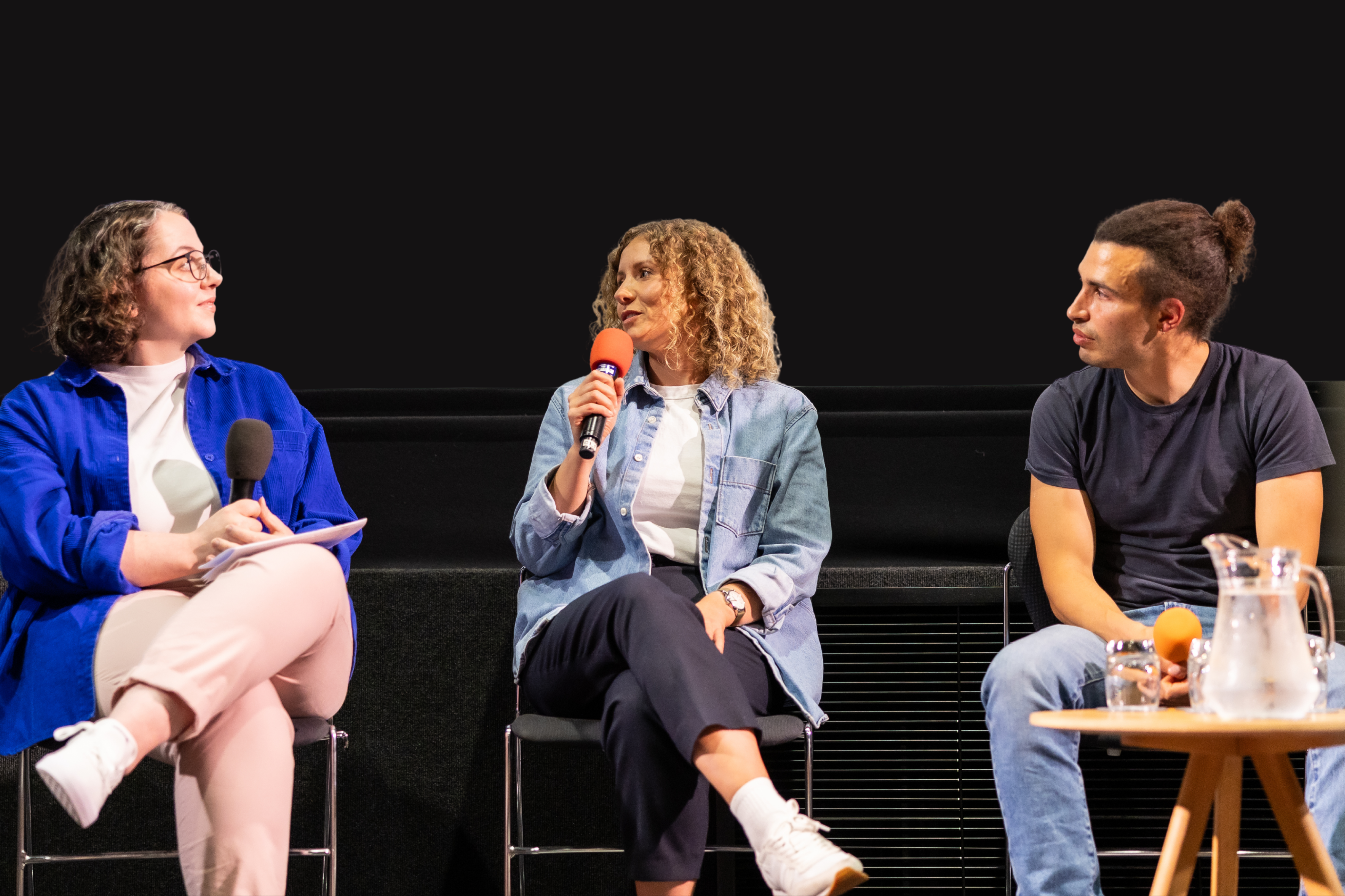 The person in the center, a woman with curly hair, is speaking into a microphone. The person on the left, wearing glasses and a blue jacket, looks towards the center speaker. The person on the right, with hair in a bun, looks to the side. A small table with a pitcher of water and glasses is in front of the person on the right.