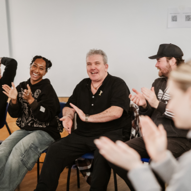 A group of people are seated in chairs, clapping and smiling, in a workshop.